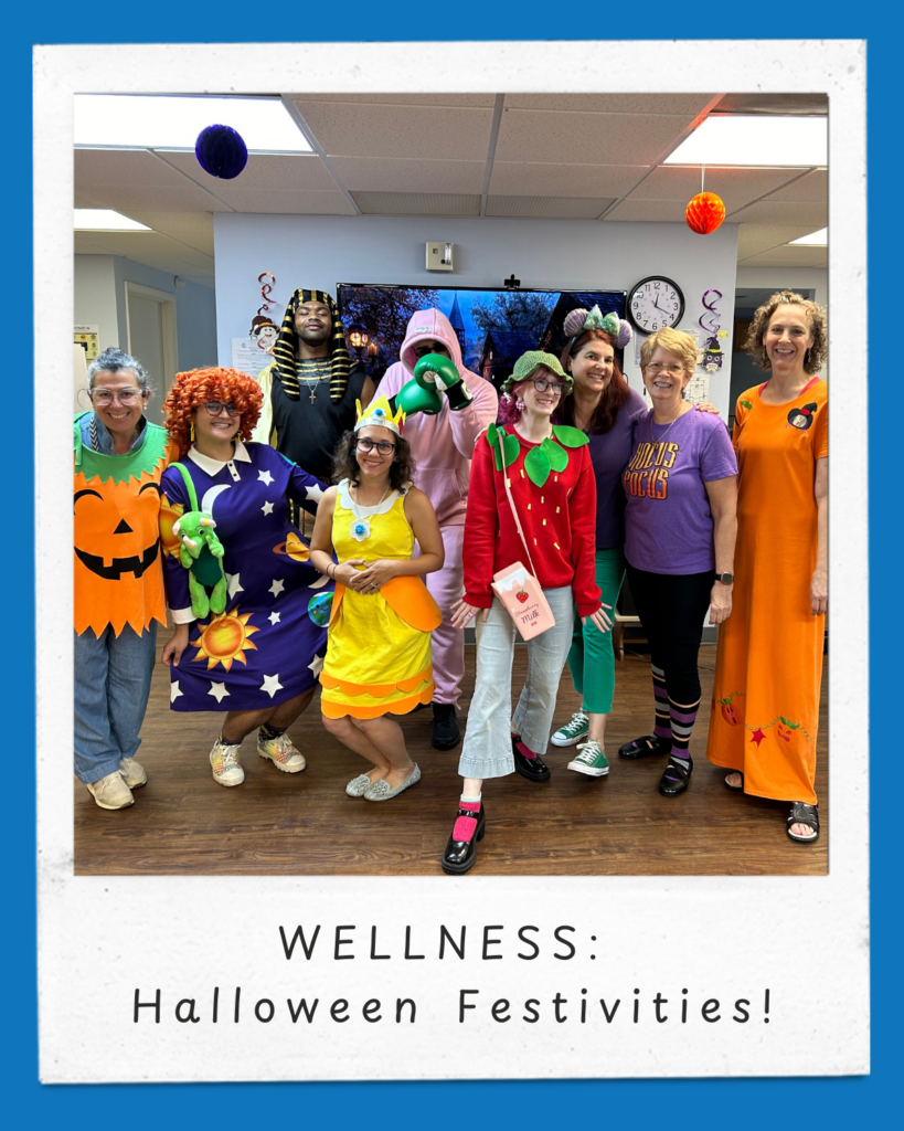 The image shows a group of nine people dressed in festive and creative Halloween costumes, standing in a bright and cheerful room decorated for the occasion. The costumes include a pumpkin, a wizard, a strawberry, a pink dinosaur, and others, showcasing a variety of vibrant colors and themes. The group is smiling and posing together, creating a fun and lively atmosphere. Behind them is a wall-mounted TV displaying a Halloween scene with trees and a darkened sky, adding to the spooky theme. The floor is wood-paneled, and there are Halloween decorations like hanging orange paper balls in the room. At the bottom of the image, there is a white frame with blue edges, featuring the text: "WELLNESS: Halloween Festivities!"