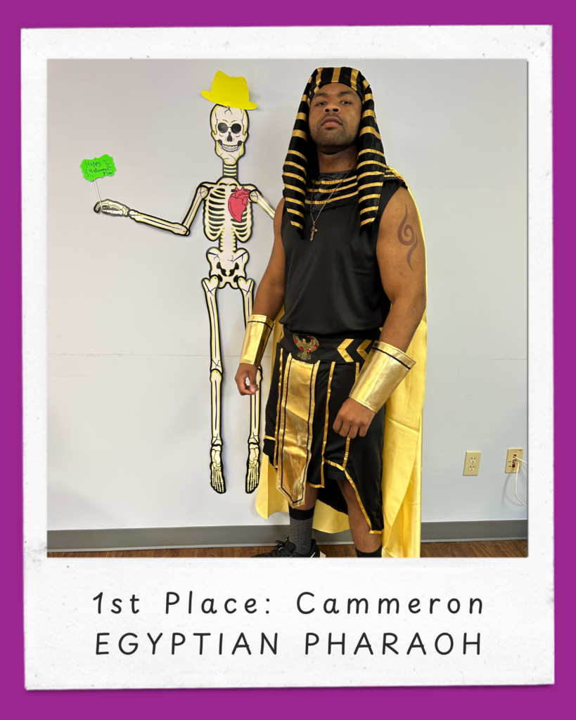 Cammeron dressed as an Egyptian Pharaoh with a black and gold costume.  He is wearing a black and gold striped headpiece and collar, black sleeveless top, black and gold bottom with belt, a gold cape, gold arm cuffs, black and grey socks and black tennis shoes. At the bottom of the image, there is a white frame with purple edges, featuring the text: "1st Place: Cammeron, Egyptian Pharaoh"