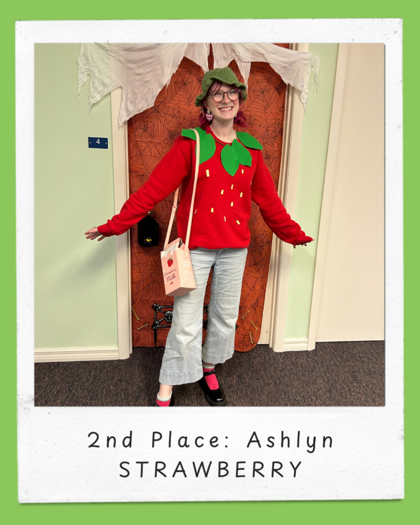 Ashlyn dressed as a Strawberry, and is wearing a red, long sleeved sweater top that has decorations of yellow seeds and a green leaf collar, a light green bucket hat, light blue pants, pink socks, black Mary Jane shoes and a purse that looks like a carton of strawberry milk. At the bottom of the image, there is a white frame with light green edges, featuring the text: "2nd Place: Ashlyn, Strawberry"