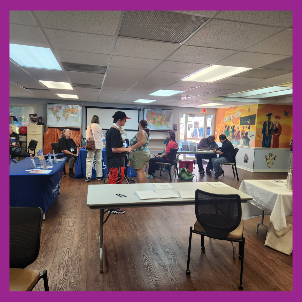 This image captures the vaccine clinic taking place in the brightly lit activity room at CIL Gainesville. Several tables and chairs are arranged throughout the room, with representatives from different organizations engaging with attendees. On the left, a table is covered with materials, while a few people are seated or interacting near it. In the center, two attendees are conversing, one holding a document or pamphlet. On the right, more people are seated at tables, some working on laptops or having discussions. The back wall features colorful maps and a projector screen, while the right wall showcases a vibrant mural with illustrations of graduates and community-themed artwork. The room has wooden flooring and a drop ceiling with fluorescent lighting. A purple border frames the image.