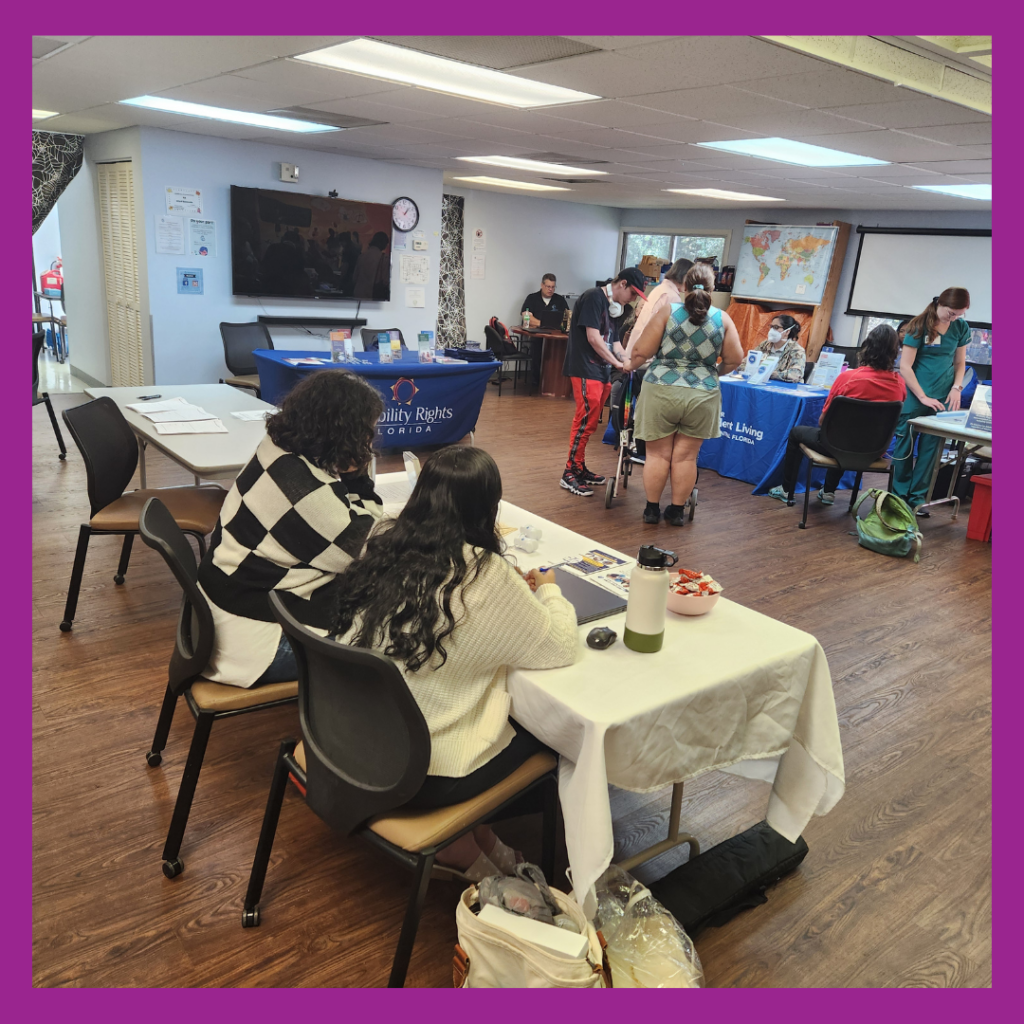 This image shows the vaccine clinic taking place in the activity room at CIL Gainesville. In the foreground, two women are seated at a table, engaged with paperwork or materials. The table is covered with a white cloth and has items like a water bottle and a bowl of snacks on it. To the right, attendees are interacting with representatives at tables for organizations, including one labeled "Disability Rights Florida" and another for the Center for Independent Living. A person using a walker is speaking with another individual near the center. The back of the room features more interactions, with tables set up displaying informational materials and a map on the wall. The room has wood flooring, a drop ceiling with fluorescent lighting, and a purple border framing the image.