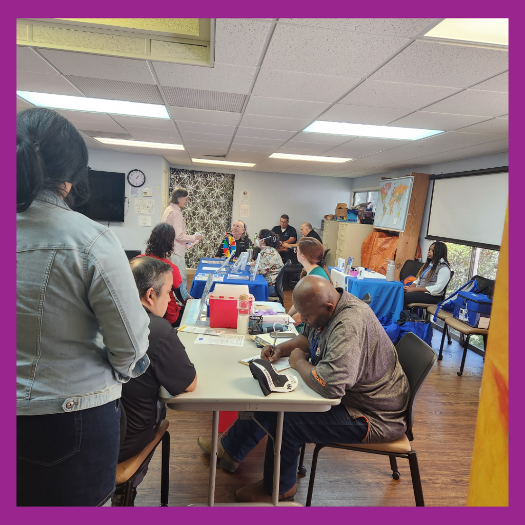 The image shows the vaccine clinic, taken place in the activity room at CIL Gainesville with several people seated at tables and engaging in various activities. The room has a casual atmosphere, with some individuals filling out forms, interacting with others, or focusing on tasks. A person standing near the center appears to be addressing the group. The tables are covered with blue tablecloths, and some have informational materials or supplies. The background includes a map, a TV screen, and a large cabinet, suggesting an organized environment. The image is bordered by a bold purple frame.