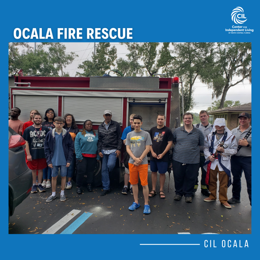 This image features a group of people standing in front of a fire truck. The group includes individuals of diverse ages and appearances, with some smiling for the camera. The fire truck is red with a closed storage compartment visible in the background. The photo is framed by a blue border with the text "OCALA FIRE RESCUE" at the top and "CIL OCALA" along the bottom-right corner, accompanied by the logo for the Center for Independent Living of North Central Florida. The setting appears to be outdoors, with trees and a cloudy sky in the background.