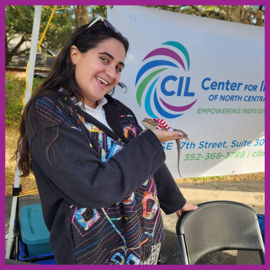 The image shows a woman (Sada) smiling and holding a bearded dragon in front of a banner for the Center for Independent Living (CIL) of North Central Florida. She is wearing a dark jacket with colorful patterns and appears happy and engaged with the pet. The bearded dragon is perched on her hand, and the banner behind her displays the CIL logo and contact information. The background includes trees and sunlight, indicating an outdoor event. The image captures a fun and friendly moment at the festival.