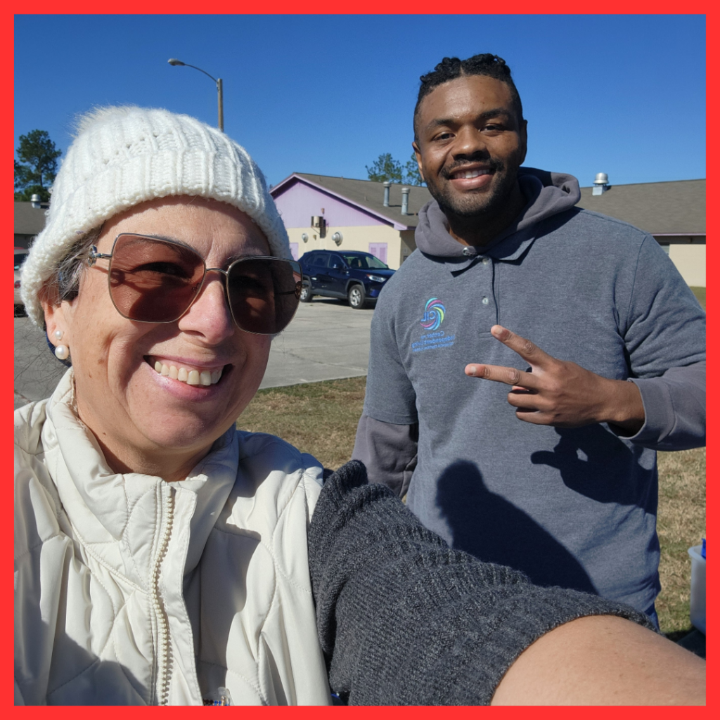 The image shows a selfie of two people standing outdoors in front of a building. On the left, a woman, Claudia, with sunglasses and a white knit hat smiles at the camera while wearing a cream-colored jacket. On the right, a man, Cammeron, with a beard and a gray shirt flashes a peace sign. The background shows a clear blue sky and a building with purple accents, and there are cars parked in the background. The red border frames the photo.