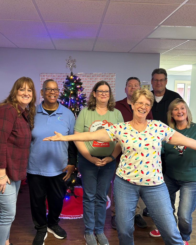 A group of seven smiling people pose in front of a decorated Christmas tree, with one person in a festive shirt playfully extending arms.