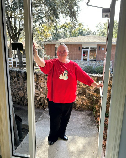 A smiling woman in a festive red snowman shirt stands outside an open door, waving and holding the handle, with a courtyard behind her.