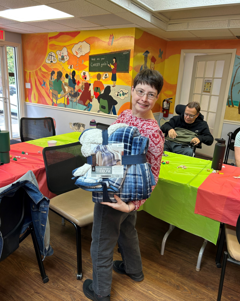 A smiling person in a red sweater holds a plush throw blanket prize in a festive room with decorated tables and a colorful mural.