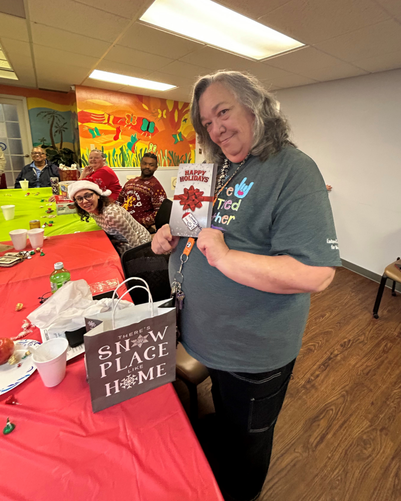 A smiling person holds a holiday card and gift bag at a festive table, with others in the background celebrating in a colorful room.