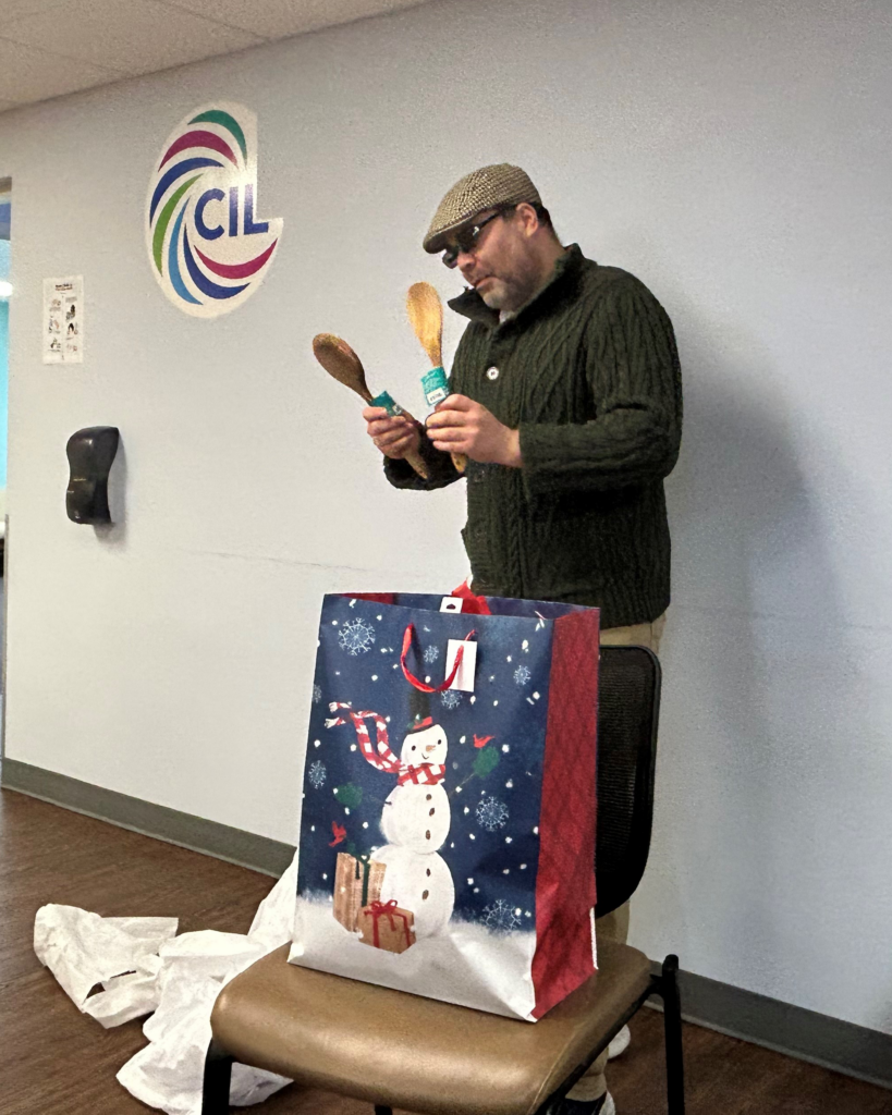 A man in a hat and sweater examines wooden spoons from a holiday gift bag with a snowman design, standing near a CIL logo on the wall.