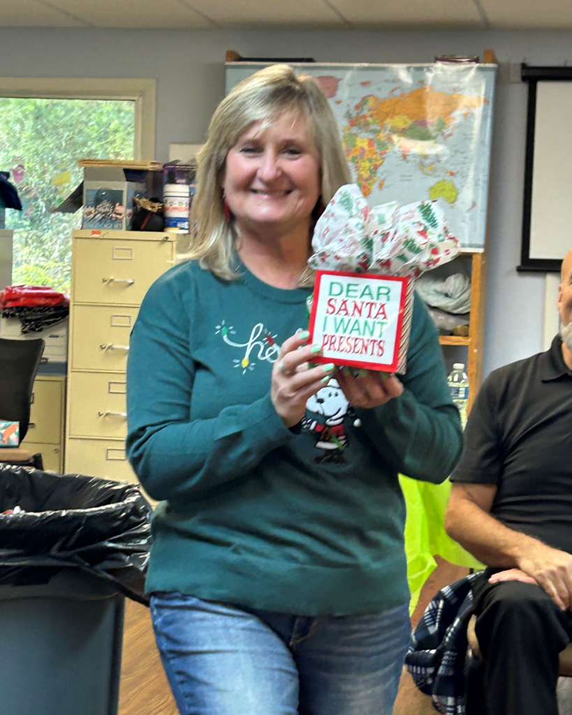 A smiling woman in a festive green sweater holds a holiday gift box that reads "Dear Santa, I Want Presents," standing in a decorated room.