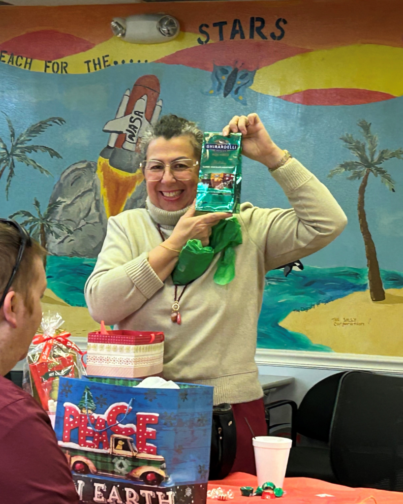 A smiling woman holds up a bag of Ghirardelli chocolates at a festive gathering, with a space-themed mural and holiday gifts behind her.