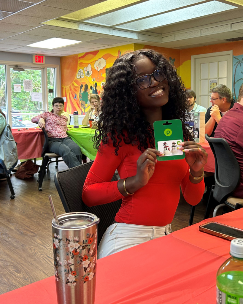 A smiling woman in a red top holds up a Publix gift card at a festive gathering, with decorated tables and a colorful mural behind her.