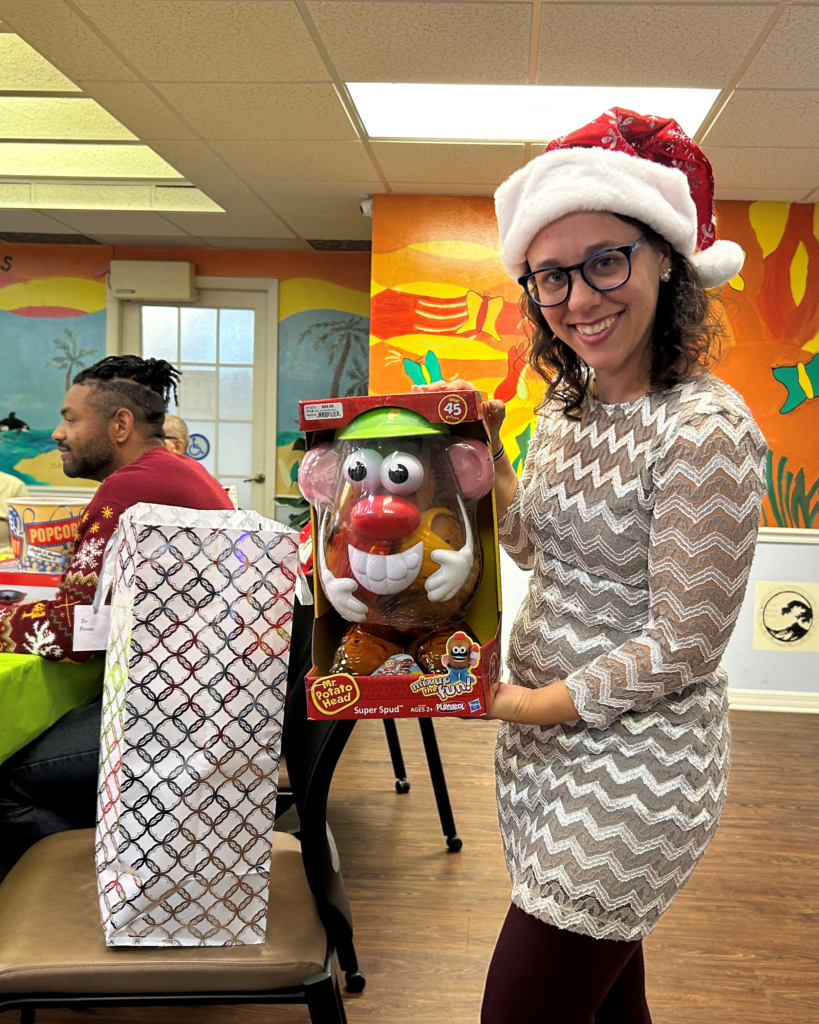 A smiling woman in a Santa hat holds a Mr. Potato Head toy at a festive gathering, with decorated tables and a colorful mural behind her.