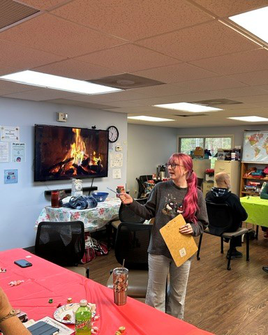 A woman with pink hair holds a folder and a small item while speaking in a festive room, with a fireplace video playing on a TV screen.