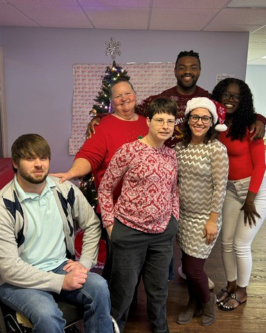 A group of six smiling people, dressed in festive attire, pose in front of a decorated Christmas tree, with one person seated in front.