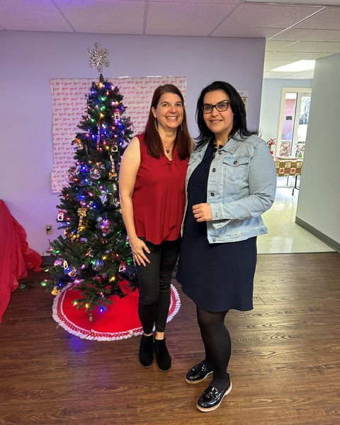 Two smiling women pose in front of a decorated Christmas tree, one wearing a red top and black pants, the other in a navy dress and denim jacket.