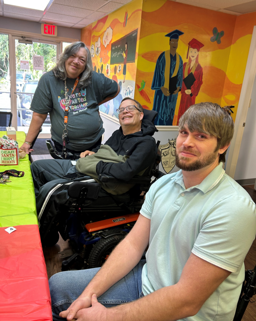 Three smiling people sit and stand around a festive table, two using wheelchairs, with a colorful mural and holiday decorations behind them.