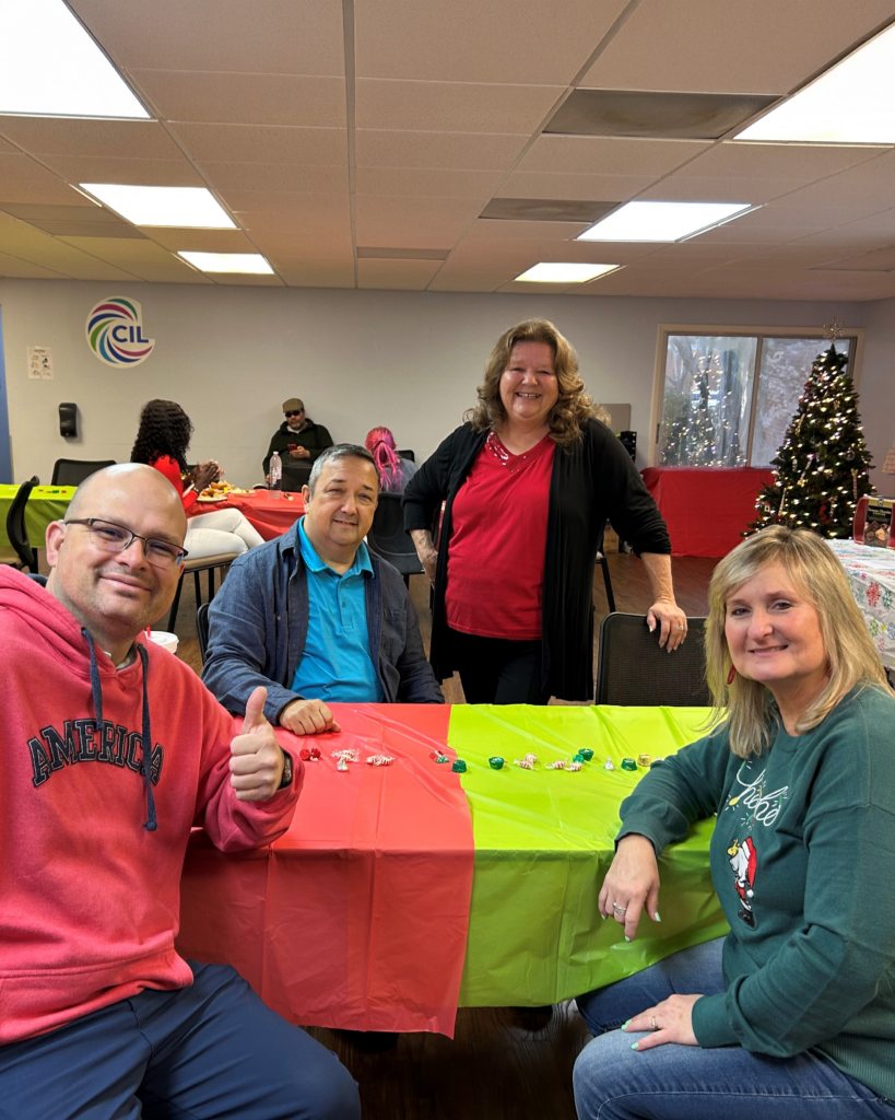 Four smiling people sit at a festive table with red and green decor, one giving a thumbs-up, with a Christmas tree in the background.