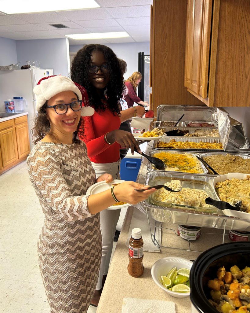 Two smiling women serve themselves from a buffet of holiday dishes, one wearing a Santa hat, in a festive gathering setting.