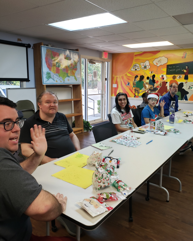 ILS participants at the Gainesville Holiday Party enjoy the festivities, sitting at a table with gift bags and crafting materials, with colorful murals in the background.