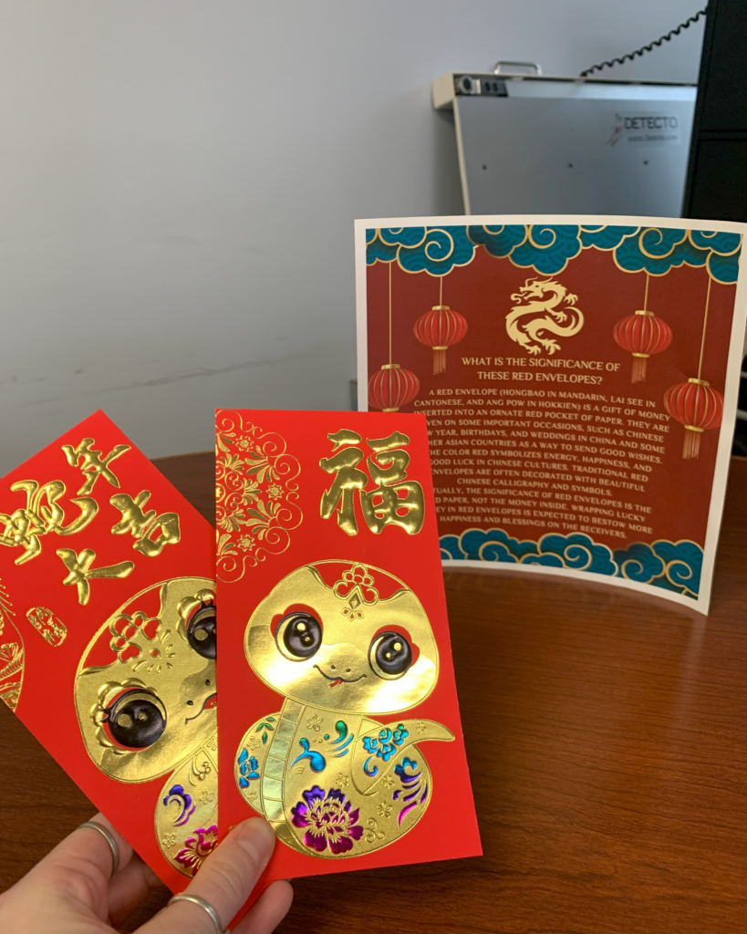 A hand holds two red envelopes with gold snake designs, in front of a sign explaining the significance of red envelopes in Lunar New Year.